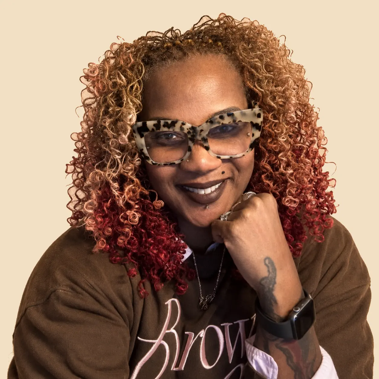 Lots of Locs owner smiling with long curled locs and glasses against the matching warm tan hero background